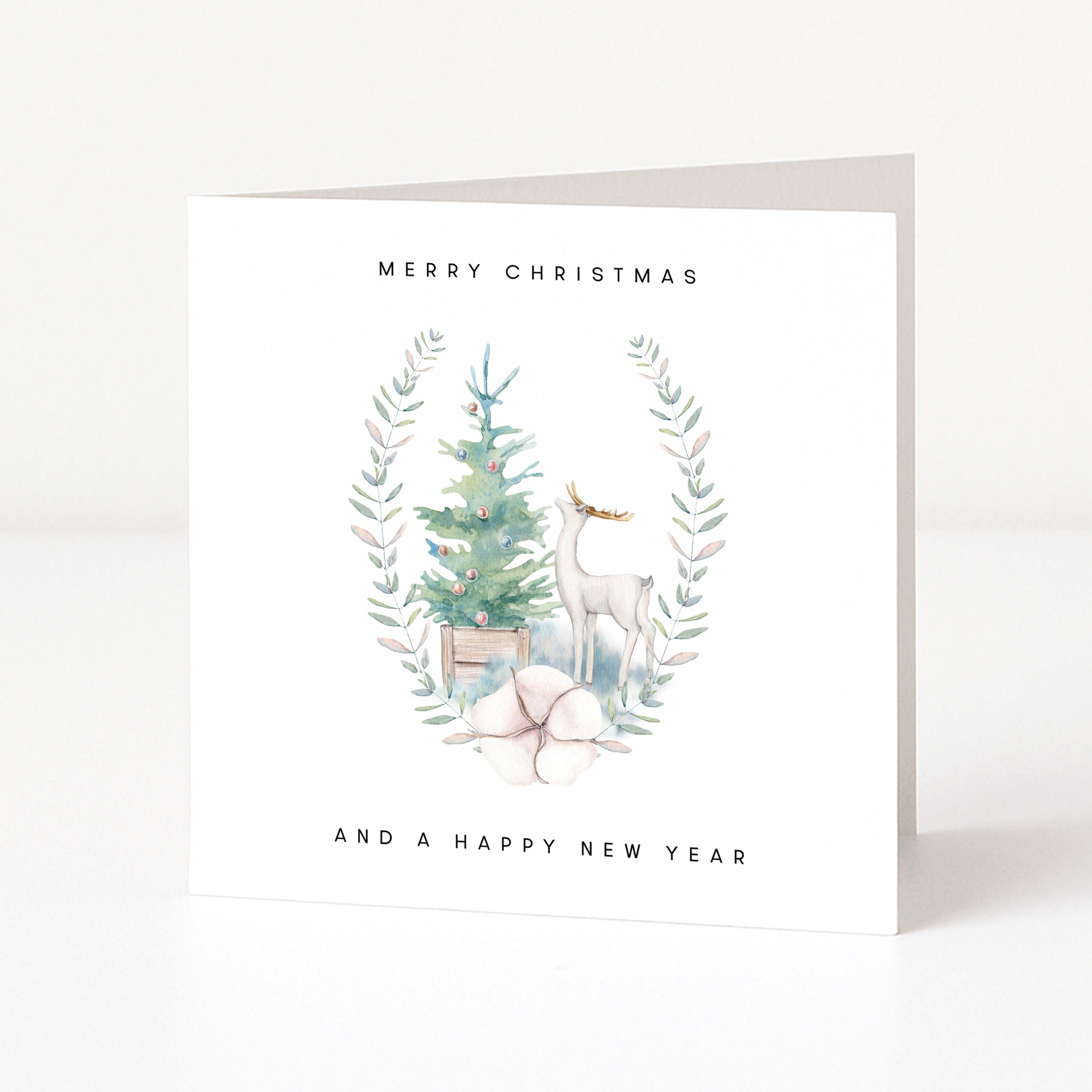 A white greeting card standing upright on a light surface, printed with the words 'MERRY CHRISTMAS AND A HAPPY NEW YEAR' around a watercolor illustration of a small tree, a deer, and pale botanical accents.