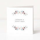 A white Christmas greeting card standing upright on a white surface, with the words 'SEASON'S GREETINGS' centered between two delicate floral garlands in soft pink, sage green, and taupe tones.