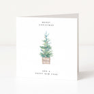 A minimalist square greeting card standing upright on a white surface. The card has a watercolour illustration of a small decorated Christmas tree in the centre, with the text 'MERRY CHRISTMAS' above and 'AND A HAPPY NEW YEAR' below.