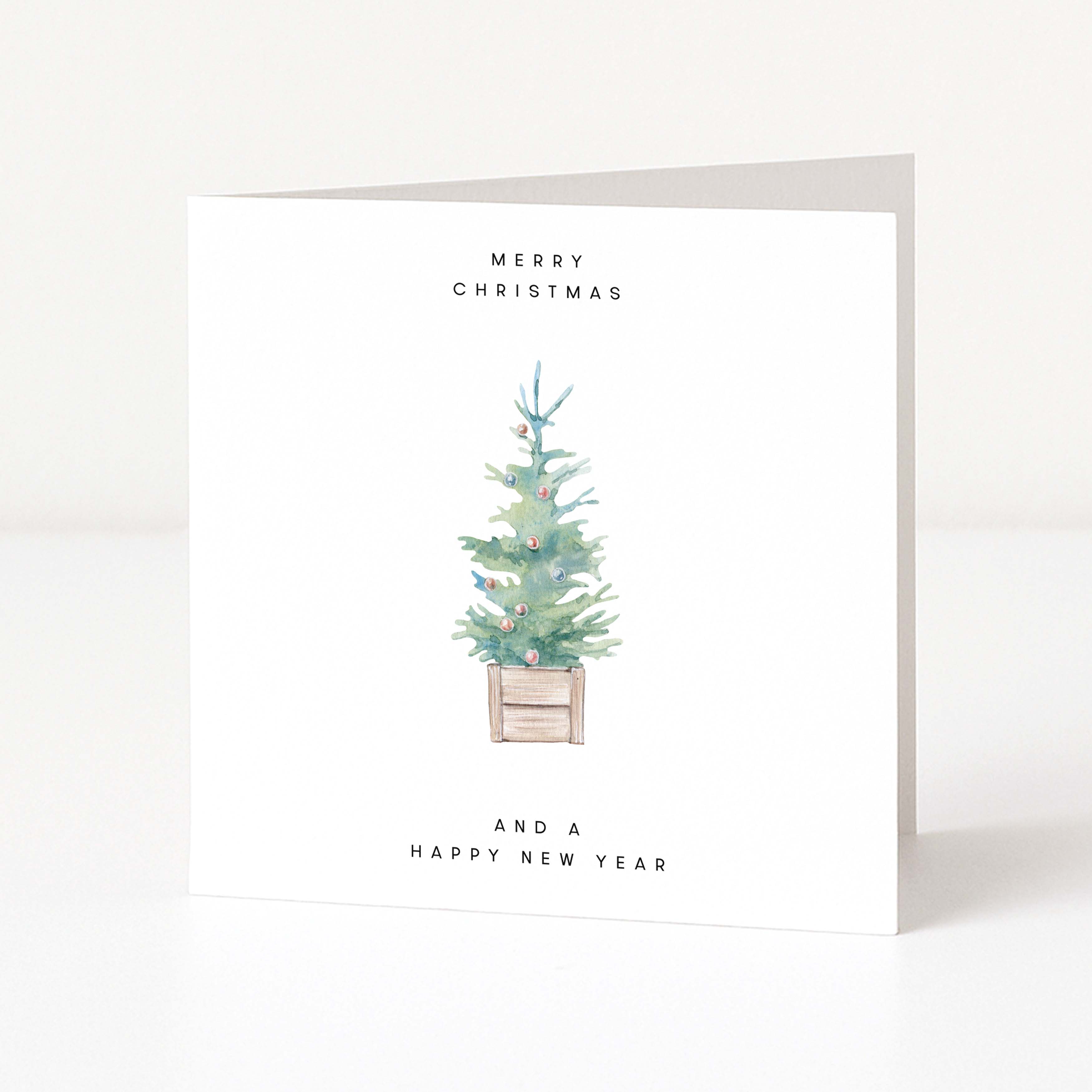 A minimalist square greeting card standing upright on a white surface. The card has a watercolour illustration of a small decorated Christmas tree in the centre, with the text 'MERRY CHRISTMAS' above and 'AND A HAPPY NEW YEAR' below.