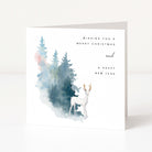 A square white greeting card standing upright on a light grey surface. The front features a watercolour-style forest with blue and green trees, a white deer with gold antlers, and the text 'Wishing you a Merry Christmas and a Happy New Year'.