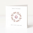 A white greeting card standing upright on a light surface, printed with the words 'MERRY CHRISTMAS' and 'AND A HAPPY NEW YEAR' around a watercolour wreath with a hanging pink ornament.