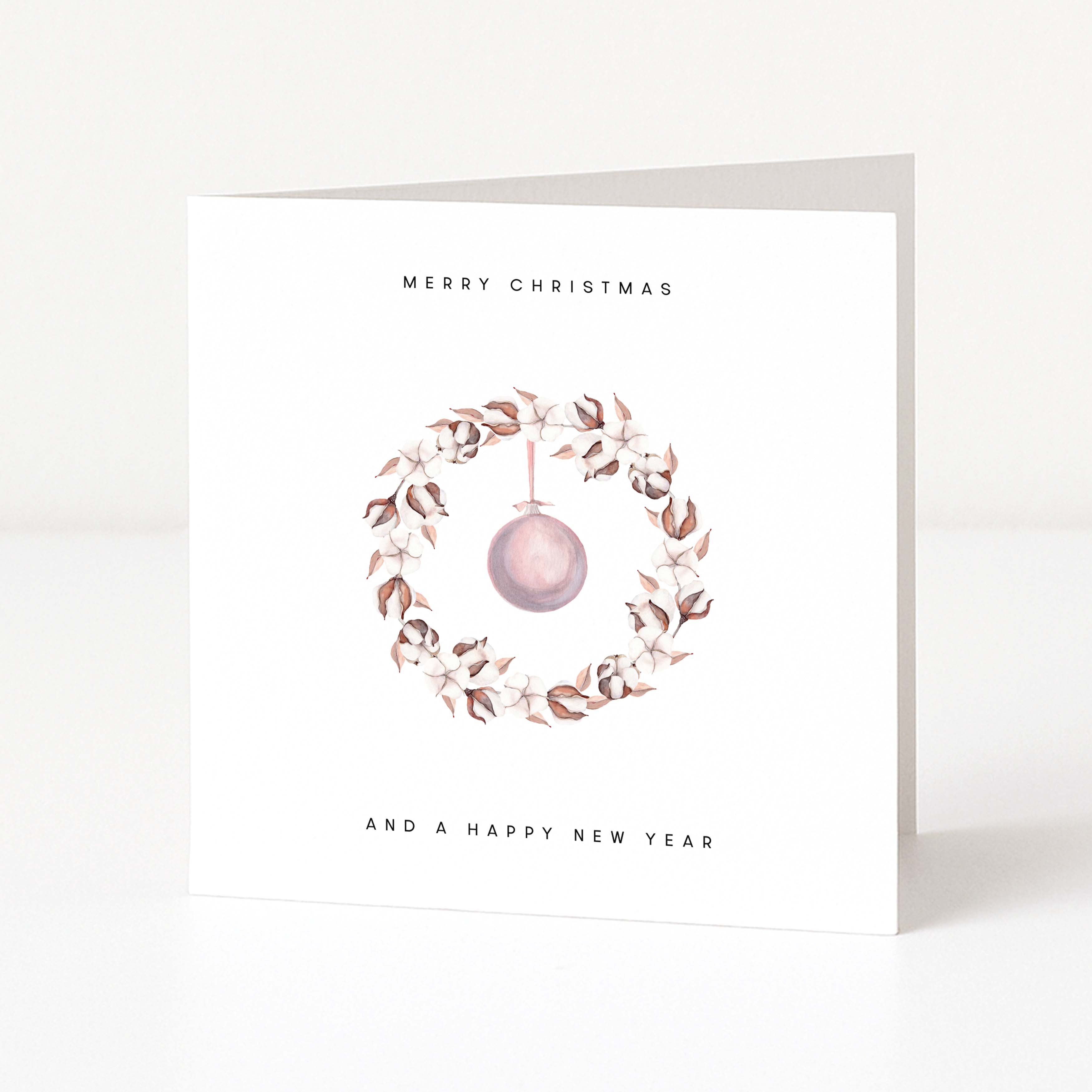 A white greeting card standing upright on a light surface, printed with the words 'MERRY CHRISTMAS' and 'AND A HAPPY NEW YEAR' around a watercolour wreath with a hanging pink ornament.
