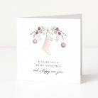 A white greeting card standing upright on a light surface, featuring a watercolour Christmas stocking decorated with pastel flowers, pinecones, and ornaments, with the text 'Wishing you a Merry Christmas and a happy new year'.