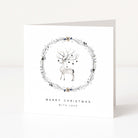 A square white Christmas greeting card standing upright on a light grey surface. The front features a watercolour-style grey deer centred within a delicate floral wreath, with the text 'MERRY CHRISTMAS WITH LOVE' printed below in black.