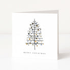 A white square Christmas greeting card standing upright on a white surface, featuring a minimalist black line drawing of a Christmas tree decorated with small metallic gold and silver ornaments, with the text 'MERRY CHRISTMAS' below.