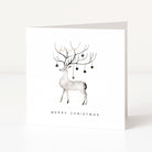 A white square Christmas greeting card standing upright on a white surface, featuring a minimalist illustration of a white deer with antlers decorated with small dark ornaments and the text 'MERRY CHRISTMAS' below.