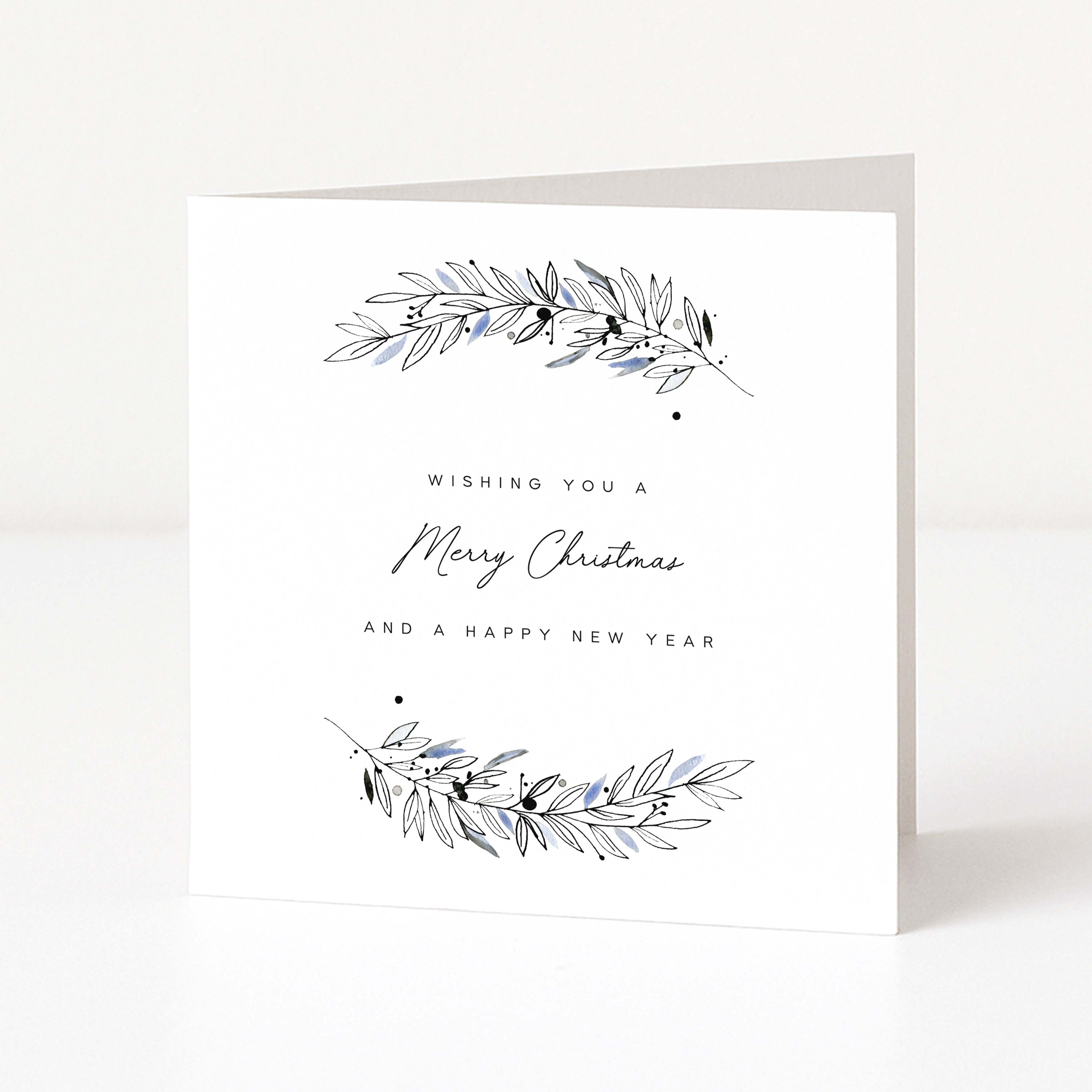 A white greeting card standing upright on a light grey surface, with delicate botanical illustrations in black and pale blue at the top and bottom and the text 'Wishing you a Merry Christmas and a Happy New Year' in the centre.