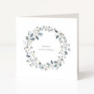 A square white Christmas greeting card standing upright, featuring a delicate wreath design in muted blue-grey and black with small gold accents, and the words “MERRY CHRISTMAS”  on the front.