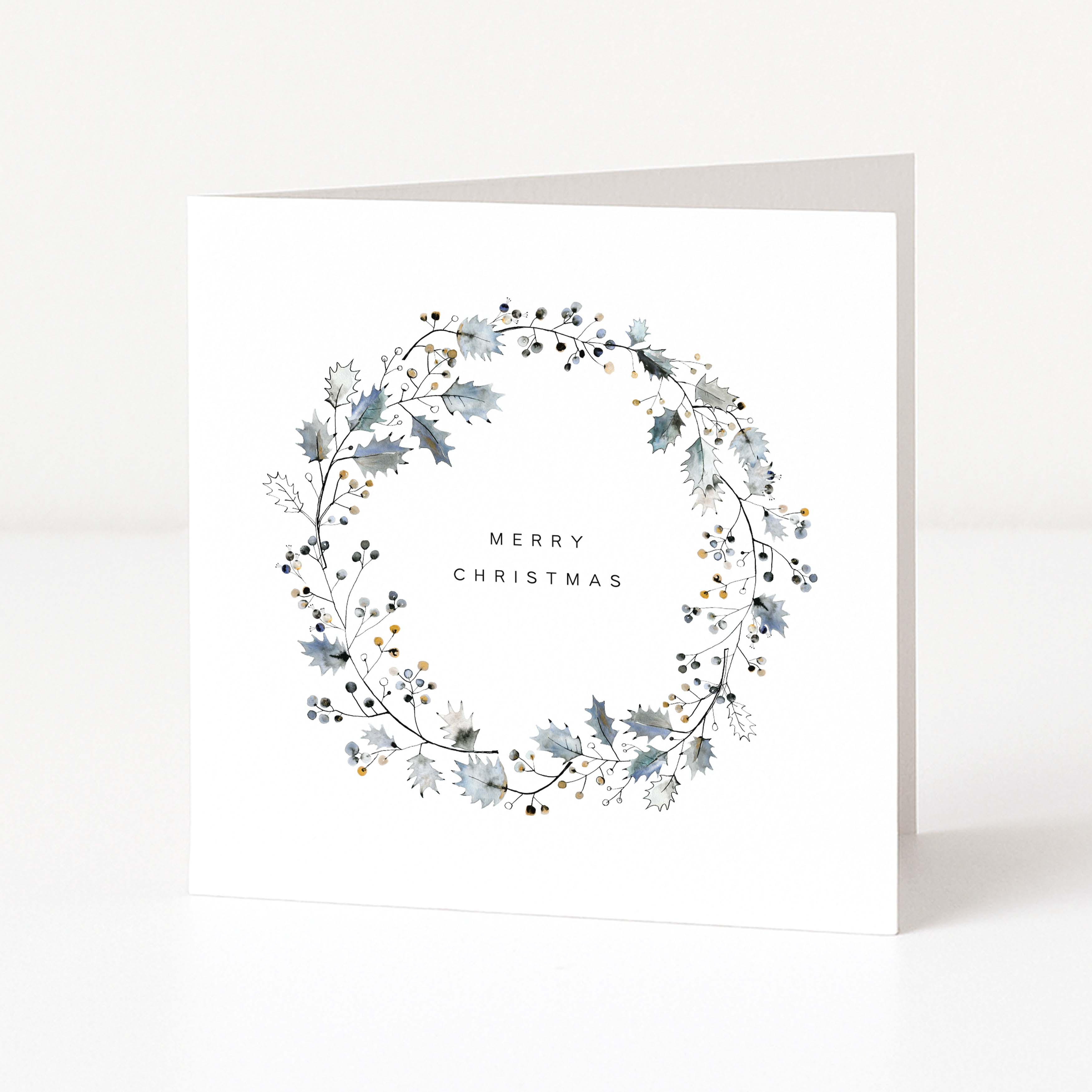 A square white Christmas greeting card standing upright, featuring a delicate wreath design in muted blue-grey and black with small gold accents, and the words “MERRY CHRISTMAS”  on the front.