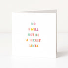 A white anti-Christmas card standing upright on a white surface, with the phrase 'NO I WILL NOT BE A SECRET SANTA' printed in small pastel letters.