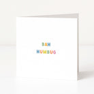A white anti-Christmas card standing upright on a white surface, with the words 'BAH HUMBUG' printed in small pastel rainbow letters near the center.