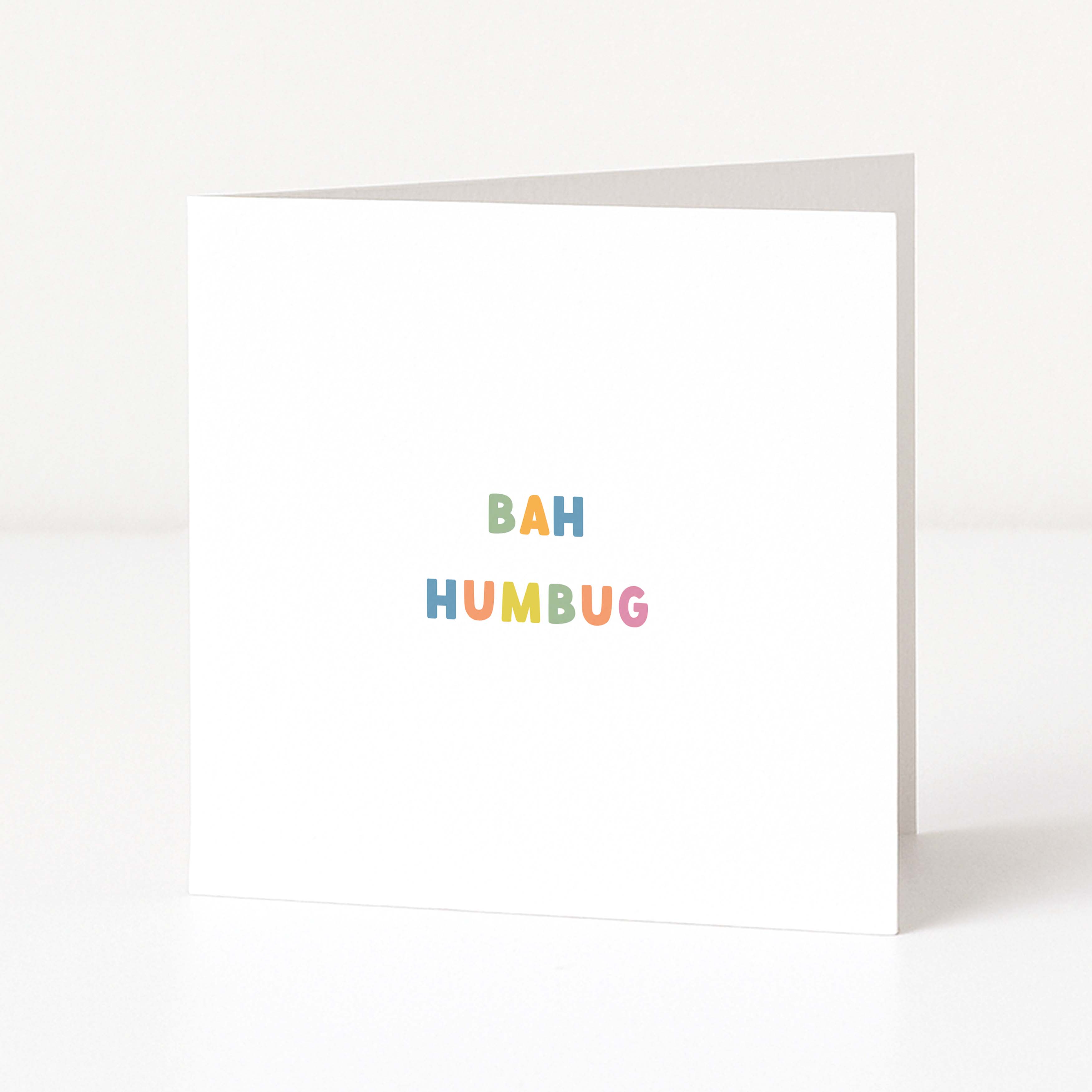 A white anti-Christmas card standing upright on a white surface, with the words 'BAH HUMBUG' printed in small pastel rainbow letters near the center.