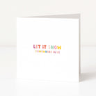 A white anti-Christmas card standing upright on a white surface, with the phrase 'LET IT SNOW (SOMEWHERE ELSE)' printed in small pastel rainbow letters near the center.