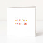 A white anti-Christmas card standing upright on a white surface, with the phrase 'ALL IS CALM ALL IS SHITE' printed in small pastel letters across the centre.