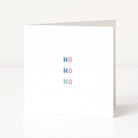 A white anti-Christmas  greeting card standing upright on a white surface, with the words 'HO HO NO' spelled out vertically in pastel blue, pink, and mint green letters.