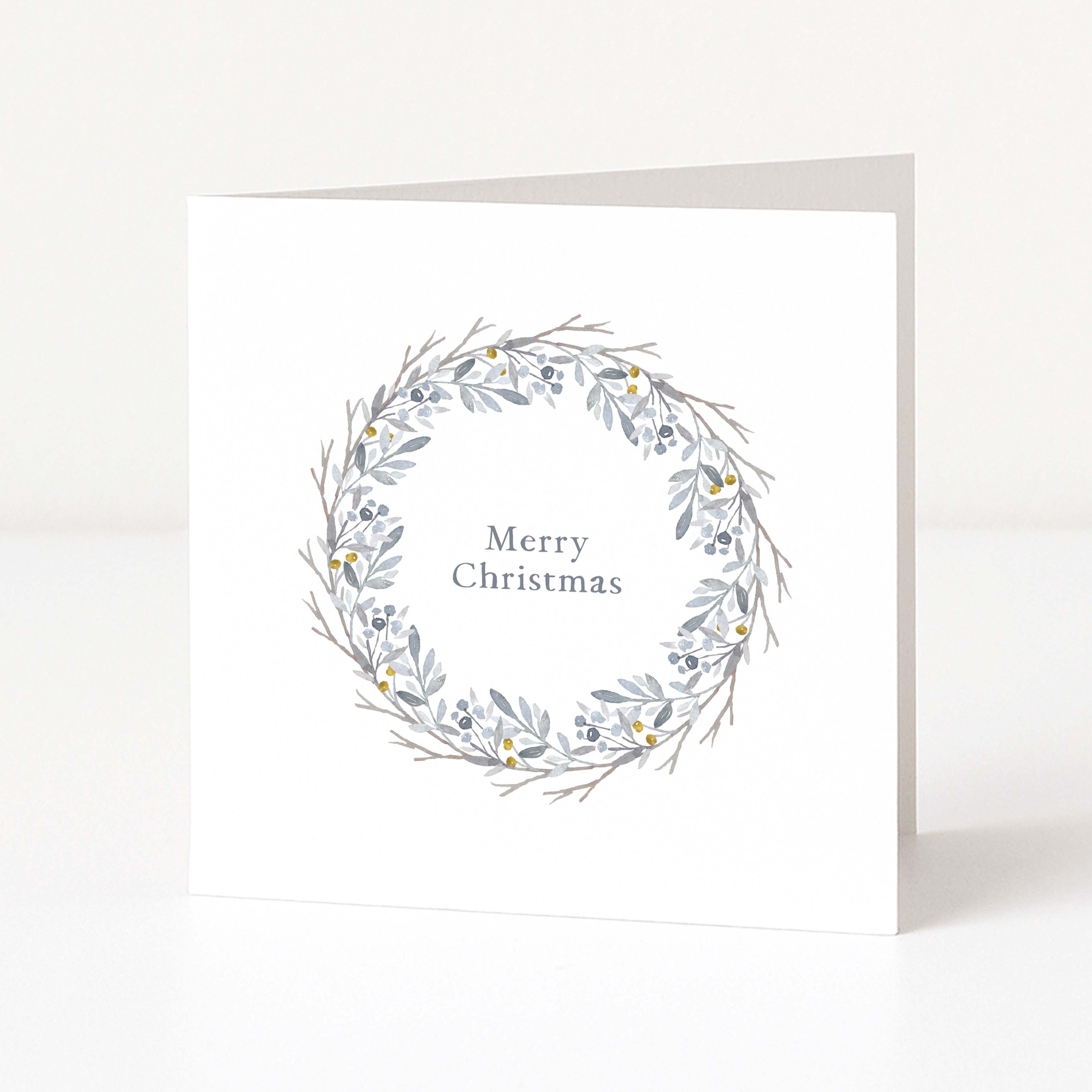 A white square Christmas greeting card standing upright on a white surface, featuring a delicate grey and blue-grey wreath illustration with small yellow accents around the words 'Merry Christmas' in a muted serif font.