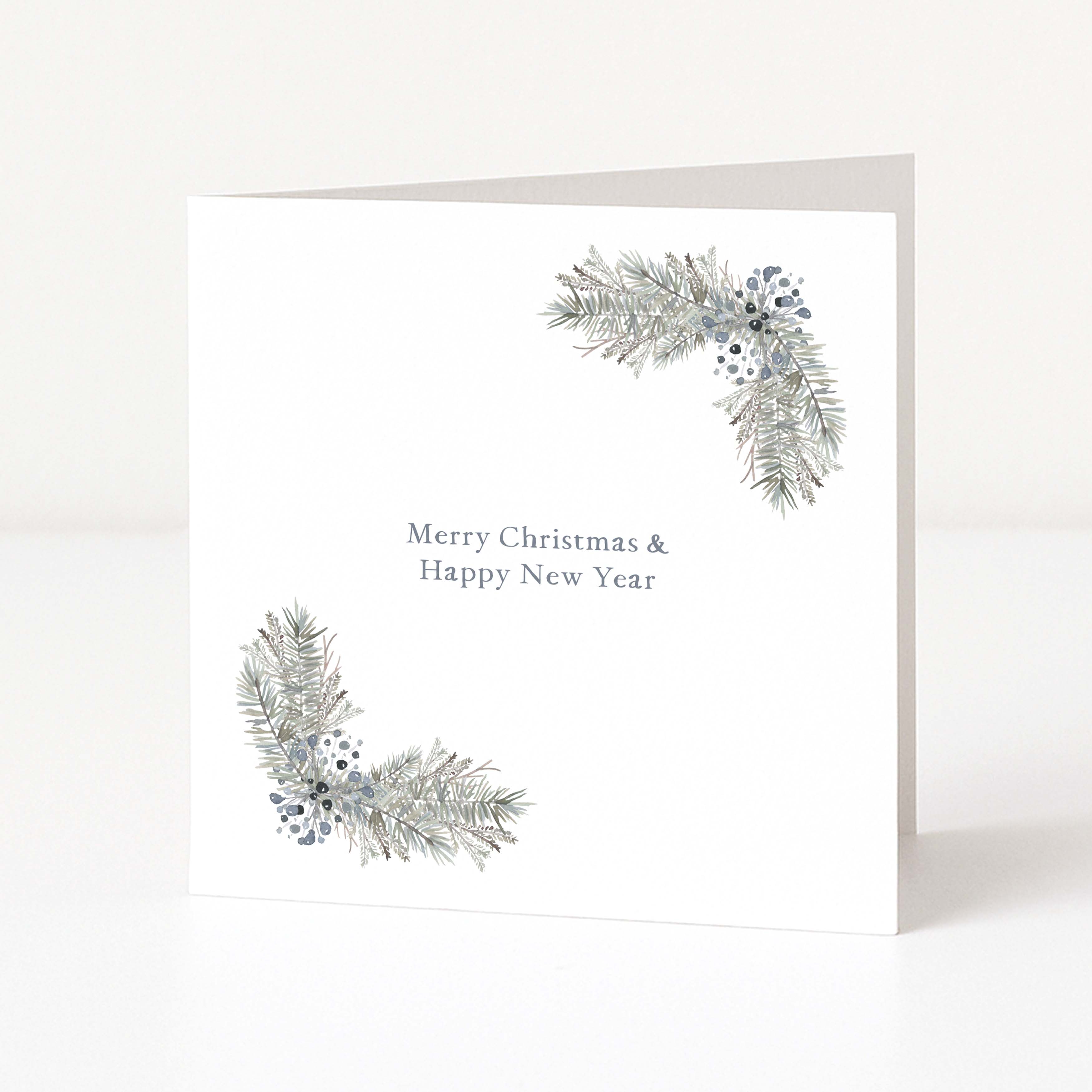 A white Christmas greeting card standing upright on a white surface, with two silver-grey pine branch corner designs and the text 'Merry Christmas & Happy New Year' in the center.