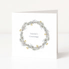 A white square Christmas greeting card standing upright on a light neutral surface. The front features a delicate watercolour wreath in muted green, grey, and gold tones with the words 'Season's Greetings' centred inside.