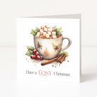 A square Christmas greeting card standing upright on a white surface. The front features an illustration of a white mug filled with hot chocolate, marshmallows, and holly leaves, with cinnamon sticks and the text 'Have a COSY Christmas' printed below.