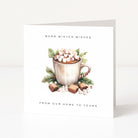 A white Christmas greeting card standing upright on a light grey surface, printed with the text 'WARM WINTER WISHES' at the top and 'FROM OUR HOME TO YOURS' at the bottom, surrounding an illustration of a mug filled with marshmallows, candy canes, greenery, and chocolates.