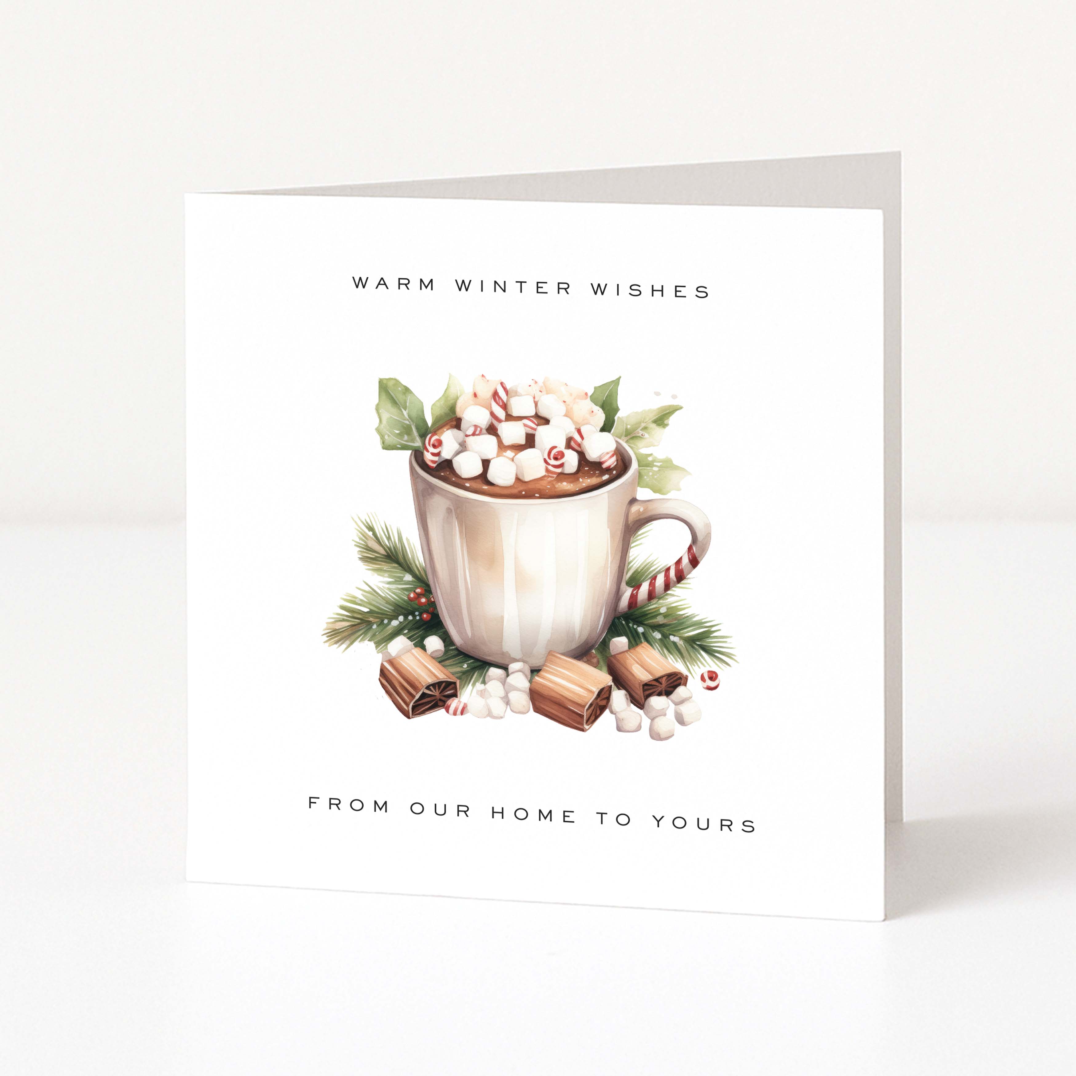 A white Christmas greeting card standing upright on a light grey surface, printed with the text 'WARM WINTER WISHES' at the top and 'FROM OUR HOME TO YOURS' at the bottom, surrounding an illustration of a mug filled with marshmallows, candy canes, greenery, and chocolates.