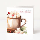 A square Christmas greeting card standing upright on a white surface against a light neutral background. The card features a watercolour illustration of a hot mug filled with marshmallows and cranberries, with the text 'SEASON'S GREETINGS' printed on the right side.