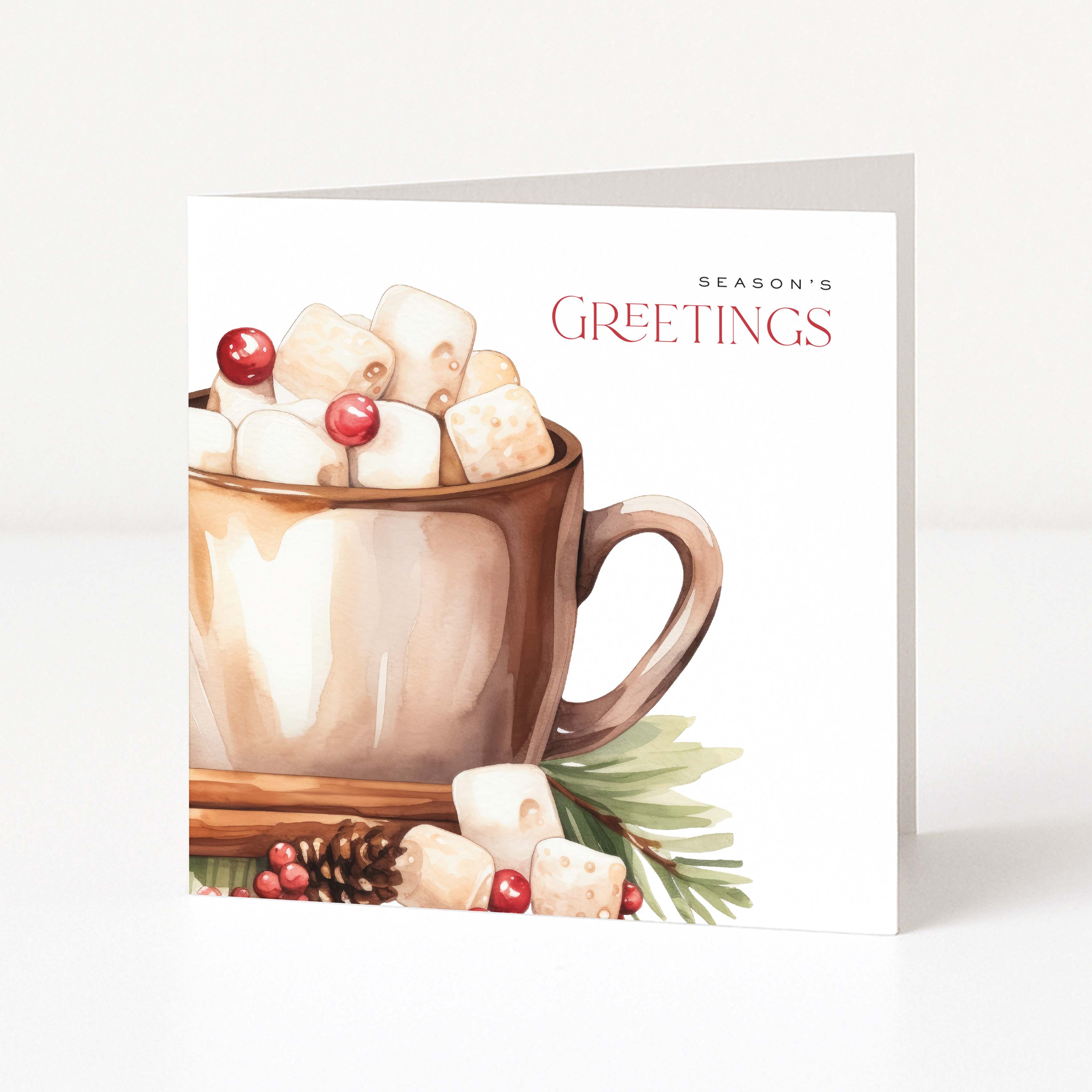 A square Christmas greeting card standing upright on a white surface against a light neutral background. The card features a watercolour illustration of a hot mug filled with marshmallows and cranberries, with the text 'SEASON'S GREETINGS' printed on the right side.