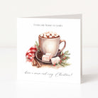 A square Christmas greeting card standing upright on a white surface. The front features an illustration of a mug filled with hot chocolate and marshmallows, with cinnamon sticks, a pinecone, and red berries, and the text 'From our home to yours' at the top and 'have a warm and cosy Christmas!' at the bottom.