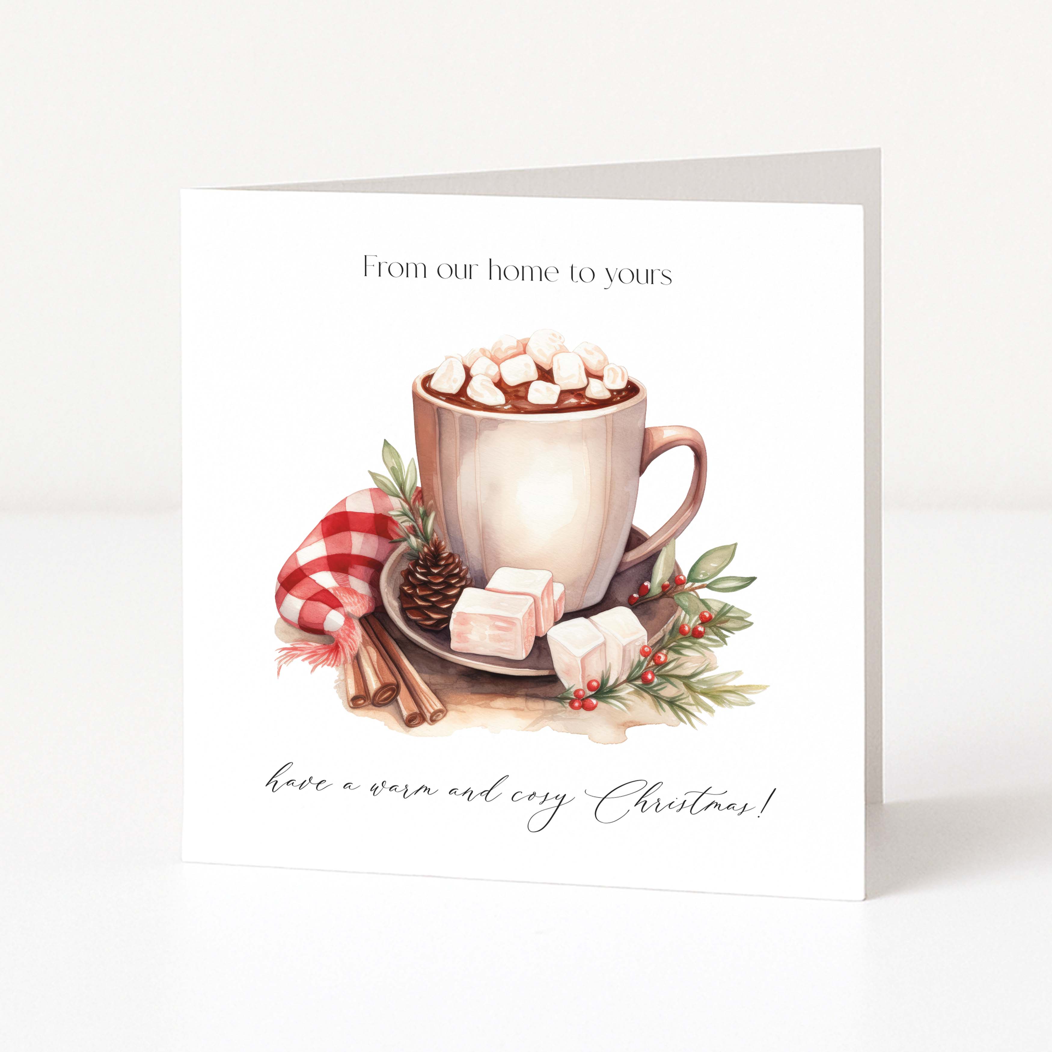 A square Christmas greeting card standing upright on a white surface. The front features an illustration of a mug filled with hot chocolate and marshmallows, with cinnamon sticks, a pinecone, and red berries, and the text 'From our home to yours' at the top and 'have a warm and cosy Christmas!' at the bottom.