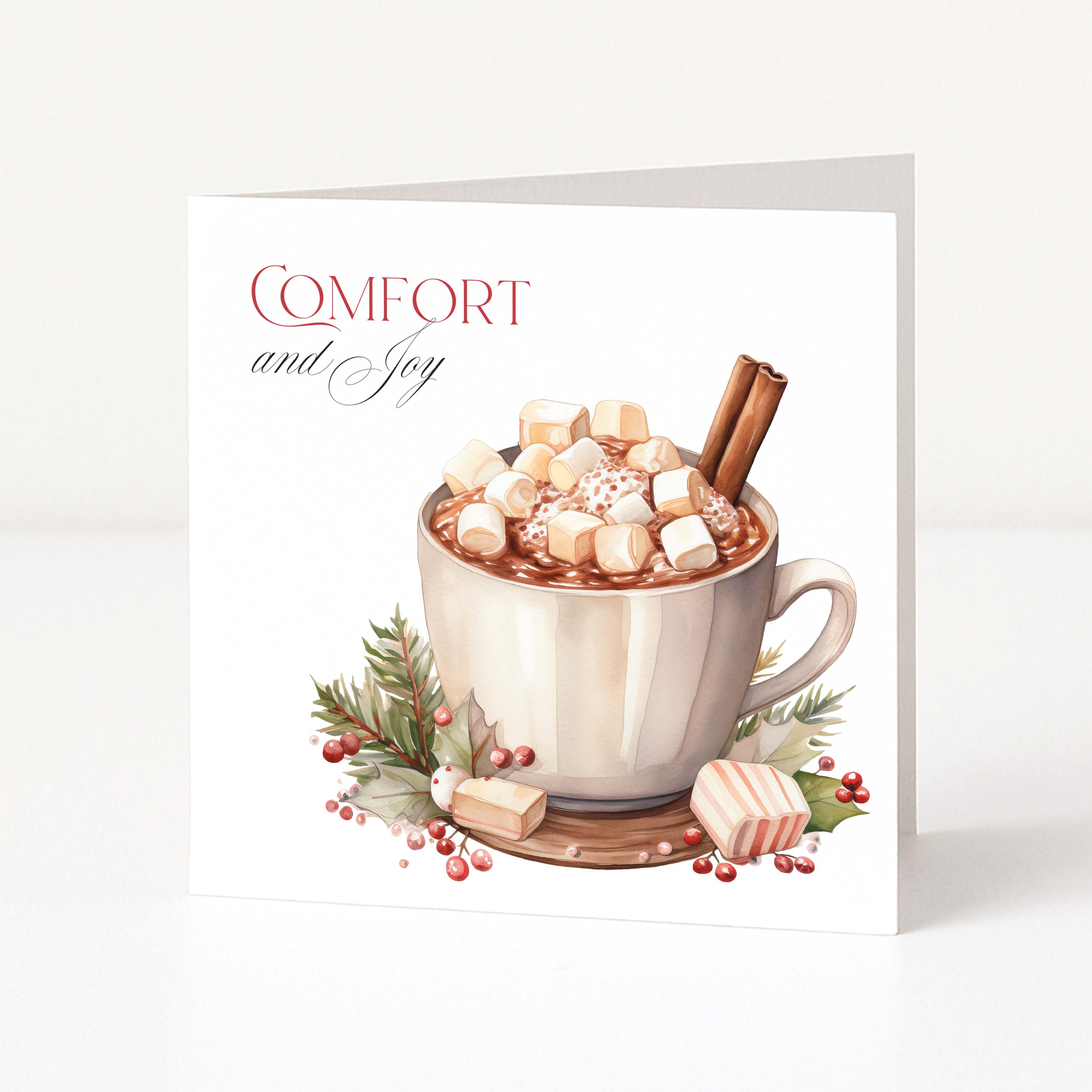 A square Christmas greeting card standing upright on a white surface, featuring a watercolour-style illustration of a mug filled with hot chocolate, marshmallows, cinnamon sticks, and holiday greenery. The card reads “COMFORT and Joy” in red and grey lettering.