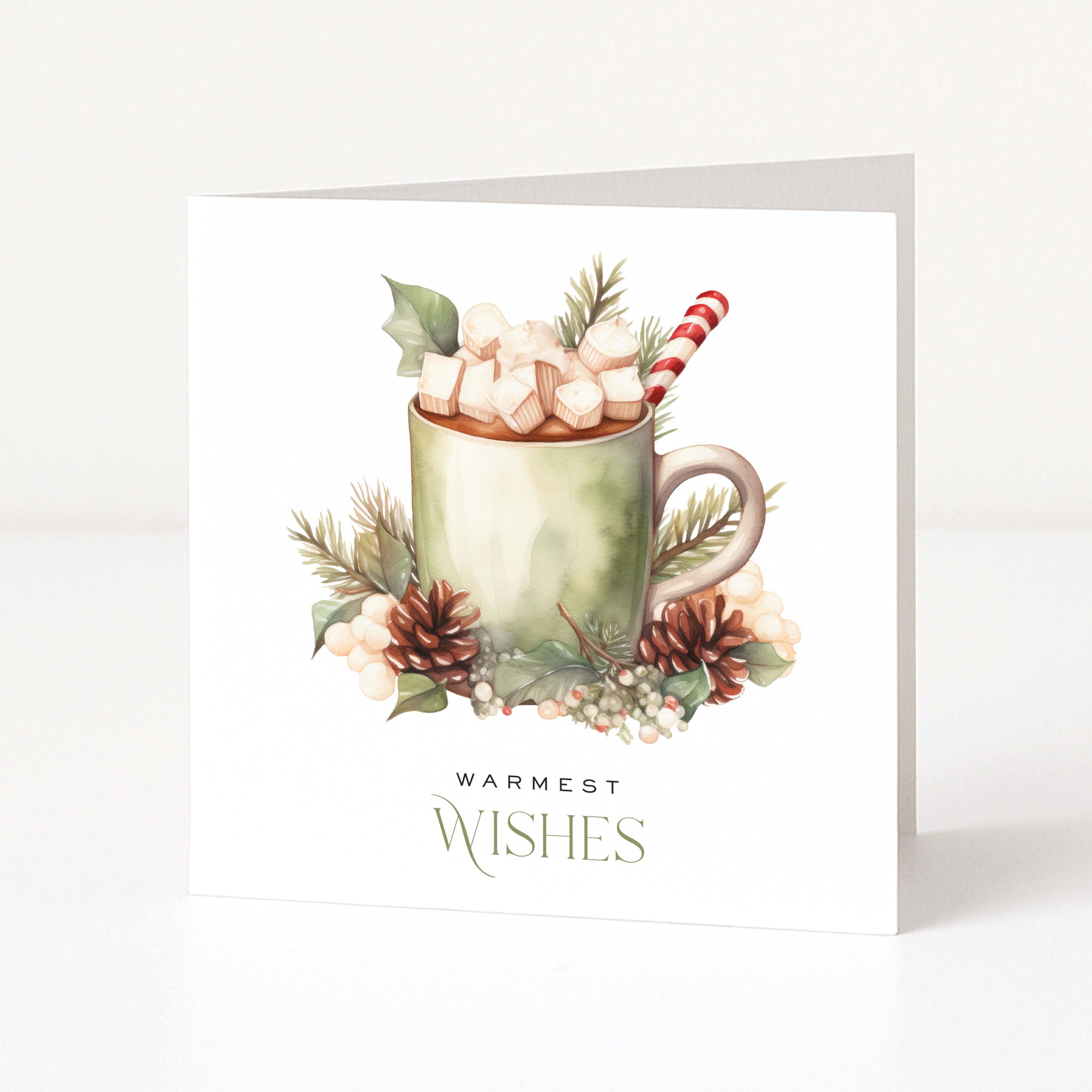 A white Christmas greeting card standing upright, featuring a watercolour illustration of a mug filled with marshmallows and a candy cane, surrounded by pinecones, berries, and greenery, with the text 'WARMEST WISHES' below.