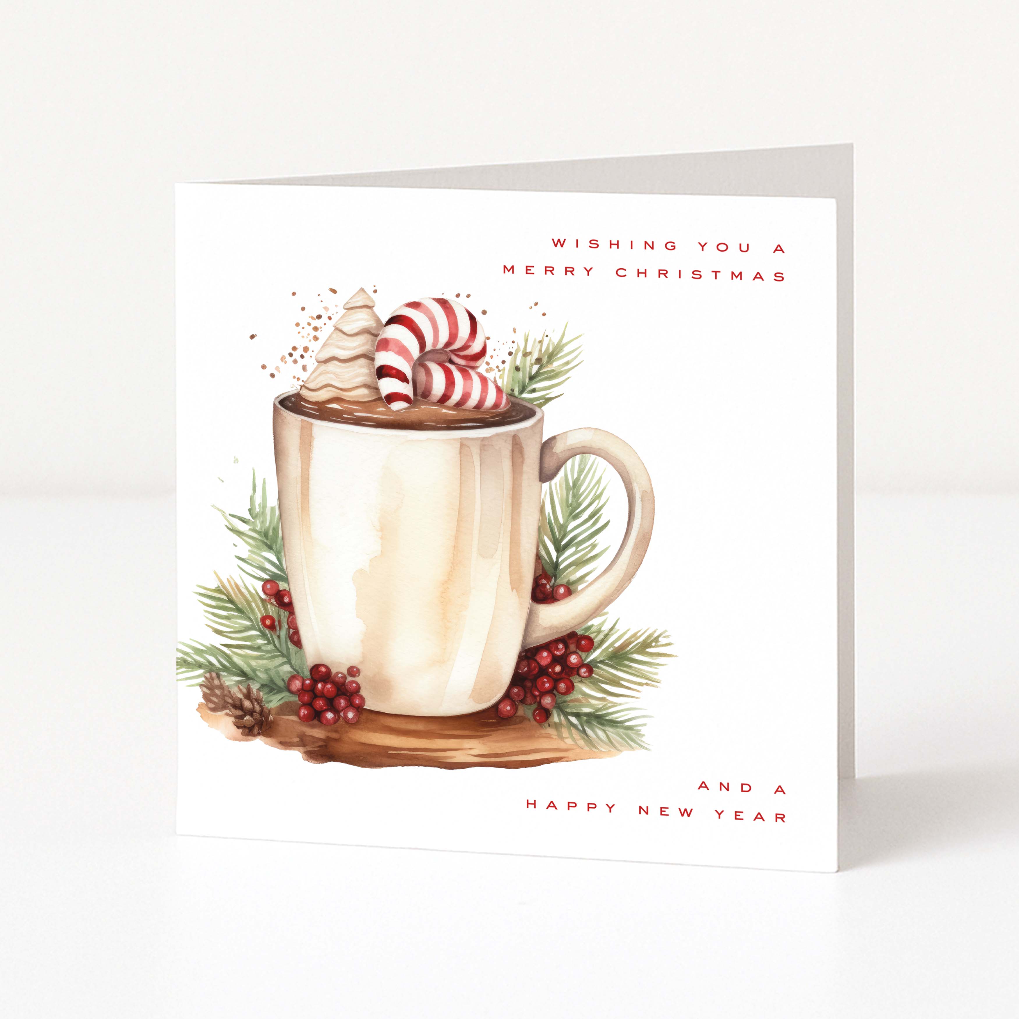 A square Christmas greeting card standing upright on a white surface. The card features a watercolor-style illustration of a beige mug filled with hot cocoa, topped with whipped cream and candy canes, surrounded by green pine branches and red berries.