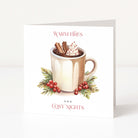 A square Christmas greeting card standing upright on a white surface against a light neutral background. The front of the card features an illustration of a mug filled with hot cocoa or coffee, topped with whipped cream and cinnamon sticks, surrounded by green leaves and red berries.
