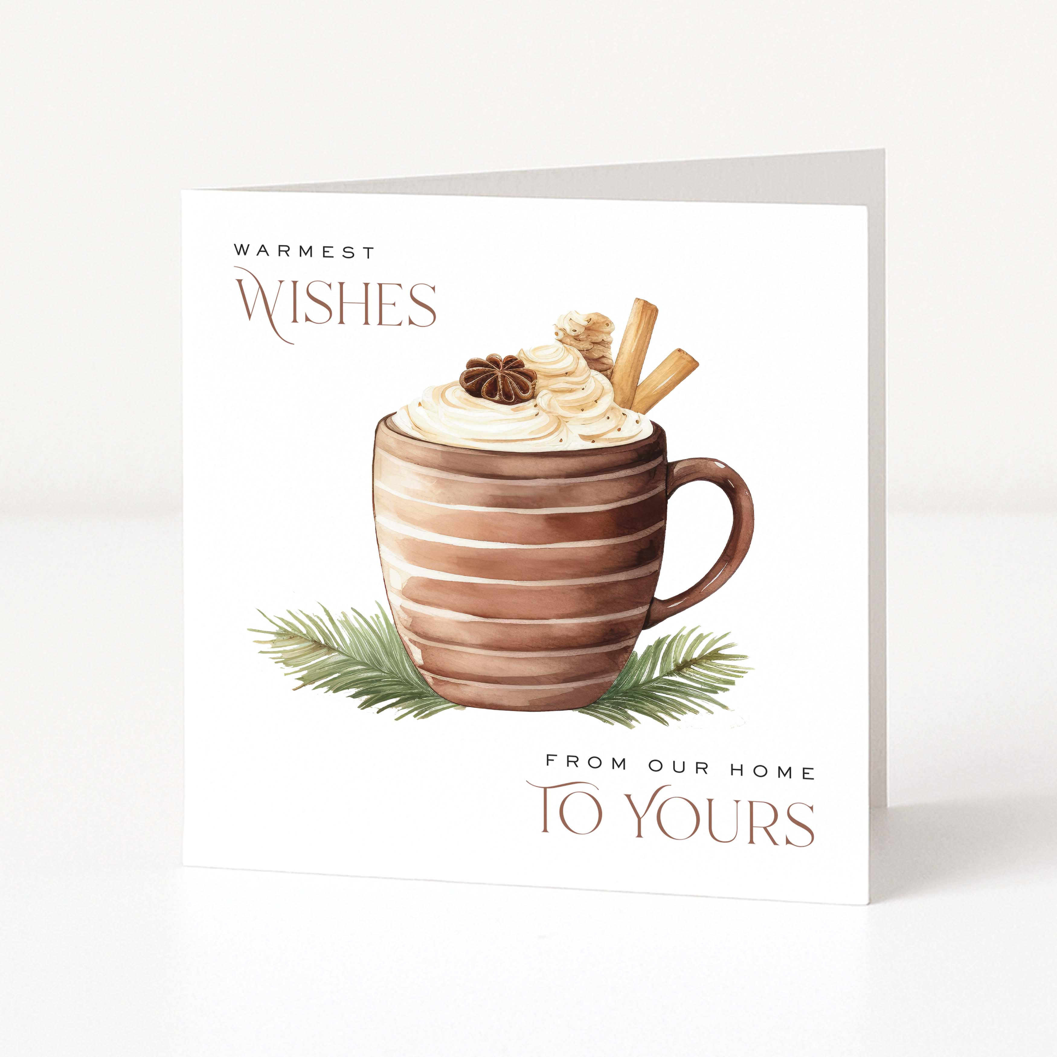 A square white Christmas greeting card standing upright on a light neutral surface. The front features an illustration of a striped brown mug filled with whipped cream and holiday toppings, with green pine branches at the base and the text 'WARMEST WISHES' and 'FROM OUR HOME TO YOURS'.