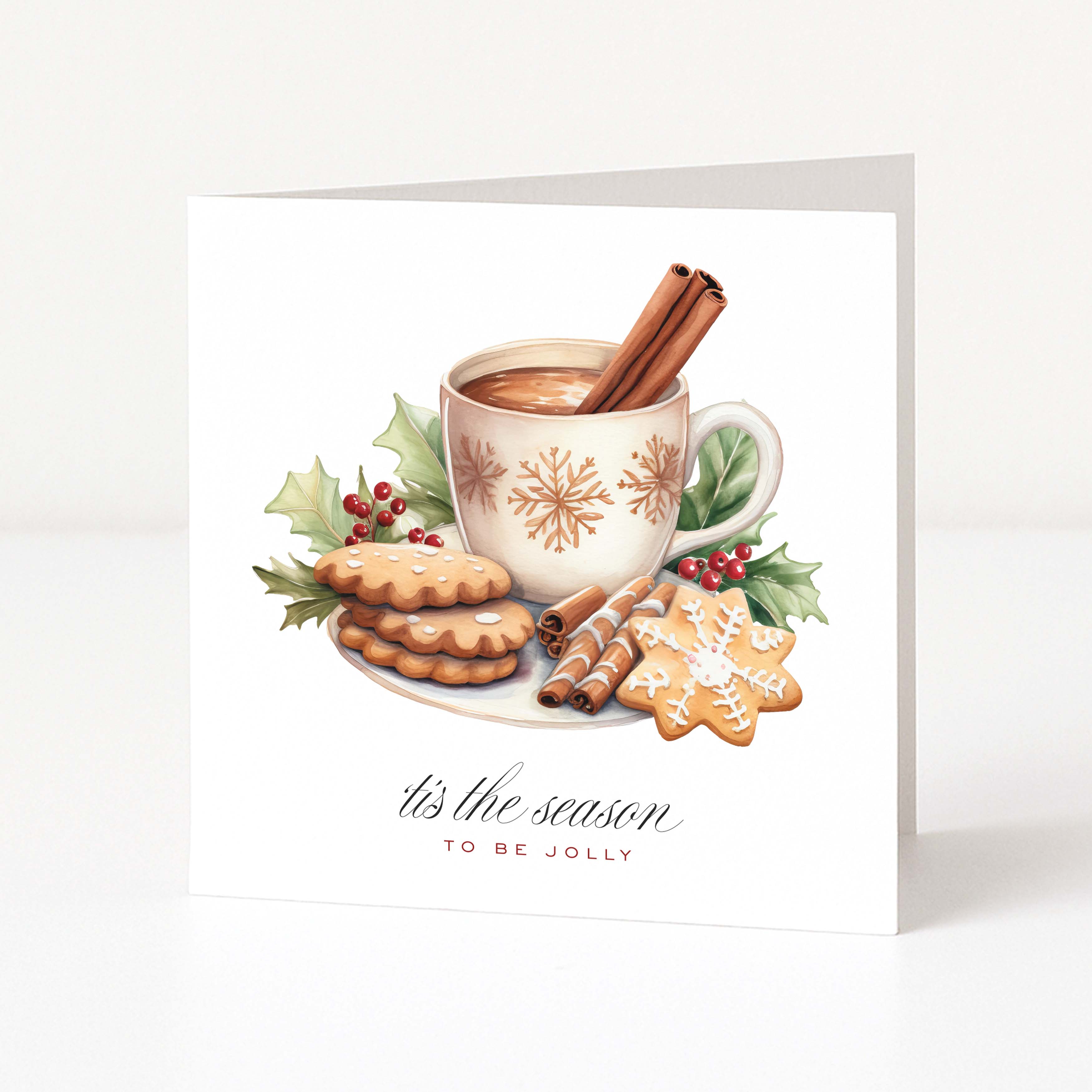 A square white Christmas greeting card with a festive illustration of a mug of hot cocoa with cinnamon sticks, cookies, holly leaves, and red berries. Below the illustration, the text reads 'it's the season TO BE JOLLY' in black and red script.