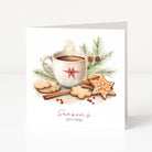 A square white Christmas greeting card standing upright, printed with a warm holiday illustration of a mug of hot cocoa, cookies, cinnamon sticks, pine branches, and the text 'Season's greetings' in red script and serif type.