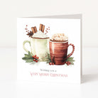 A square Christmas greeting card standing upright on a white surface. The card features an illustration of two hot cocoa mugs topped with whipped cream, with cinnamon sticks, star anise, holly leaves, and red berries.