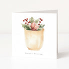 A square white greeting card standing upright on a white surface, printed with a watercolor illustration of holly leaves, red berries, and a striped ornament in a beige cup, with the text 'Season's Greetings' below.
