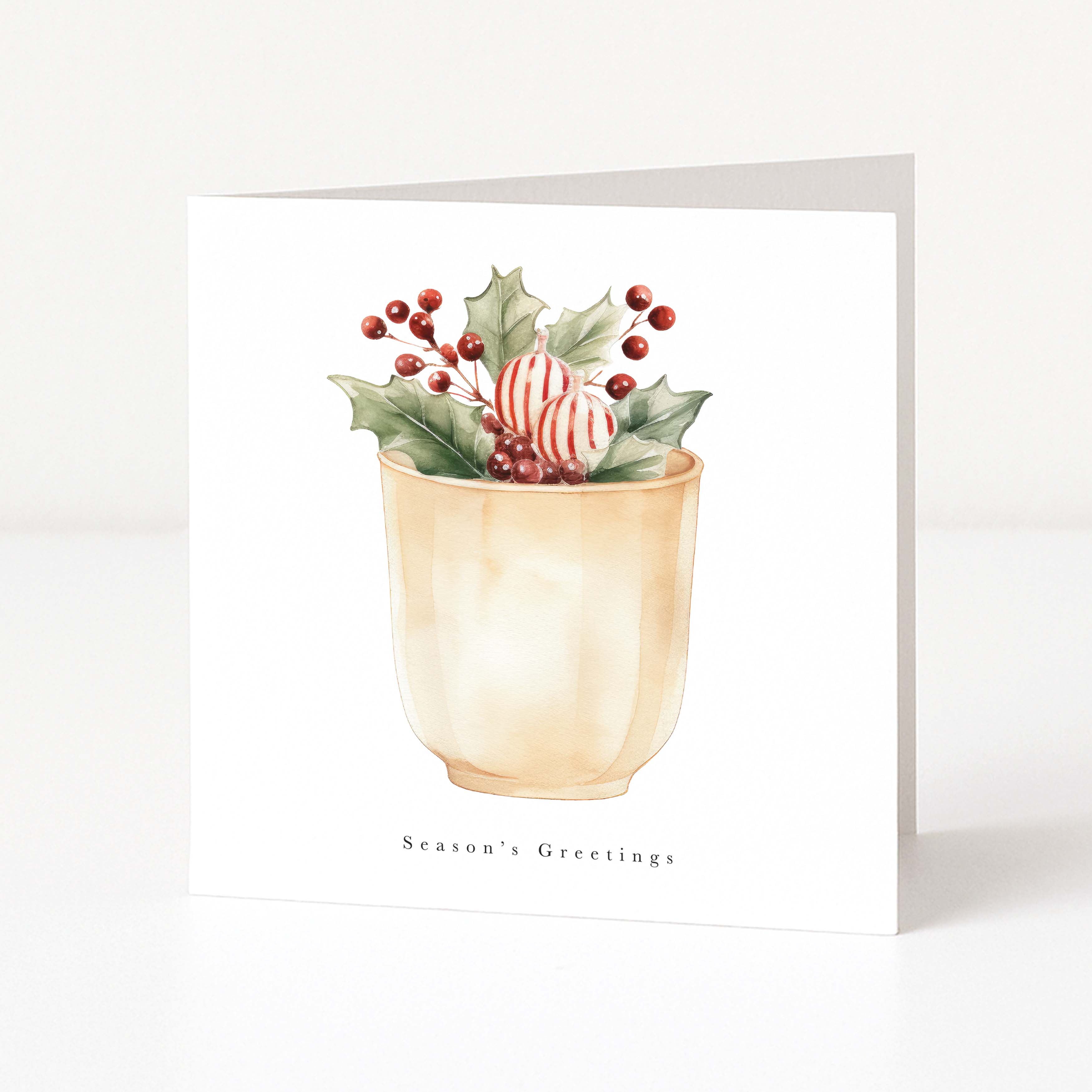 A square white greeting card standing upright on a white surface, printed with a watercolor illustration of holly leaves, red berries, and a striped ornament in a beige cup, with the text 'Season's Greetings' below.