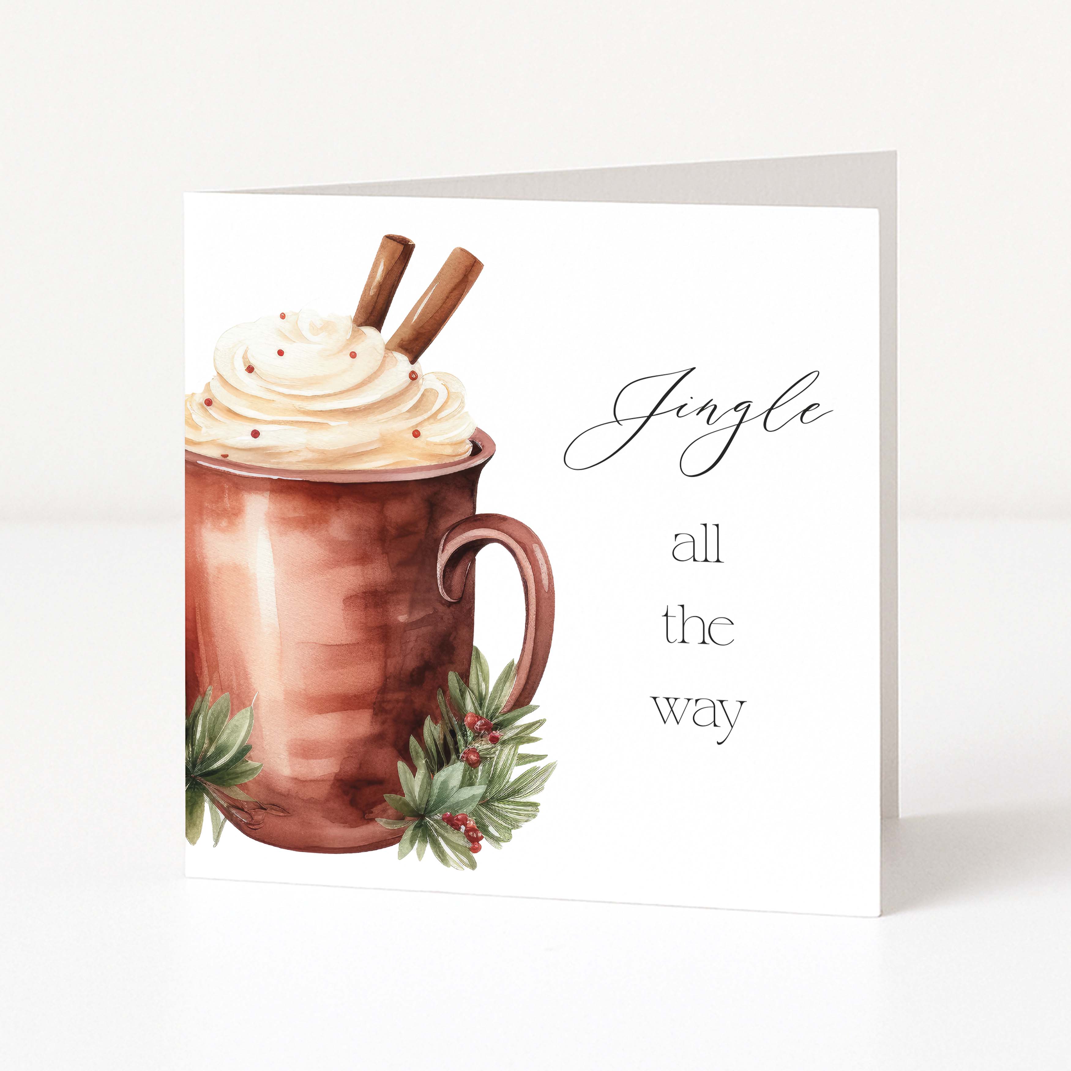 A white Christmas greeting card with a watercolor illustration of a copper mug filled with whipped cream, cinnamon sticks, and holly, next to the text 'Jingle all the way'.