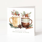 A square white Christmas greeting card with a watercolor illustration of two holiday mugs topped with whipped cream, holly, pinecones, and red berries. The card reads 'Season's Greetings FROM OUR HOME TO YOURS' and is photographed standing upright on a clean white surface against a white background.