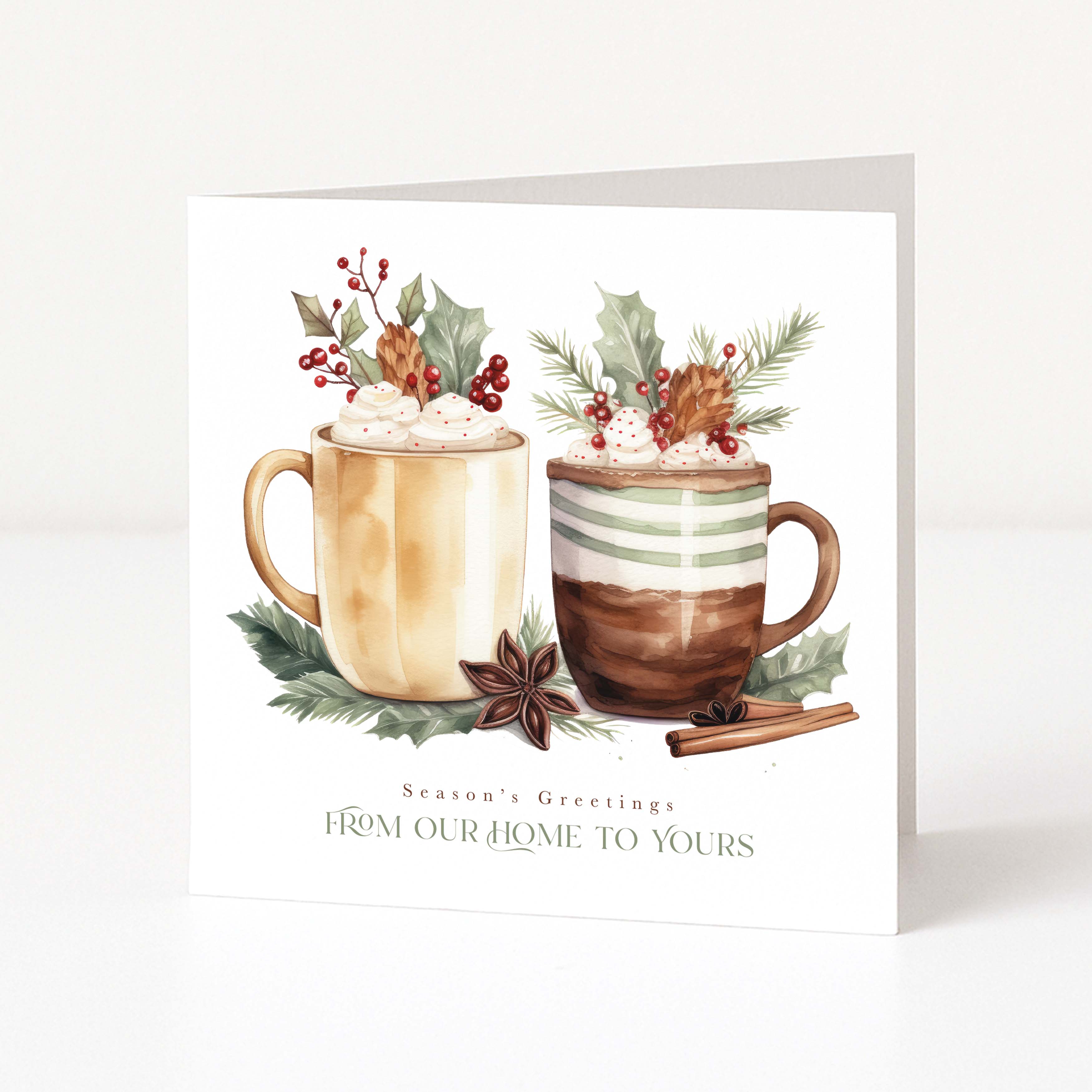 A square white Christmas greeting card with a watercolor illustration of two holiday mugs topped with whipped cream, holly, pinecones, and red berries. The card reads 'Season's Greetings FROM OUR HOME TO YOURS' and is photographed standing upright on a clean white surface against a white background.