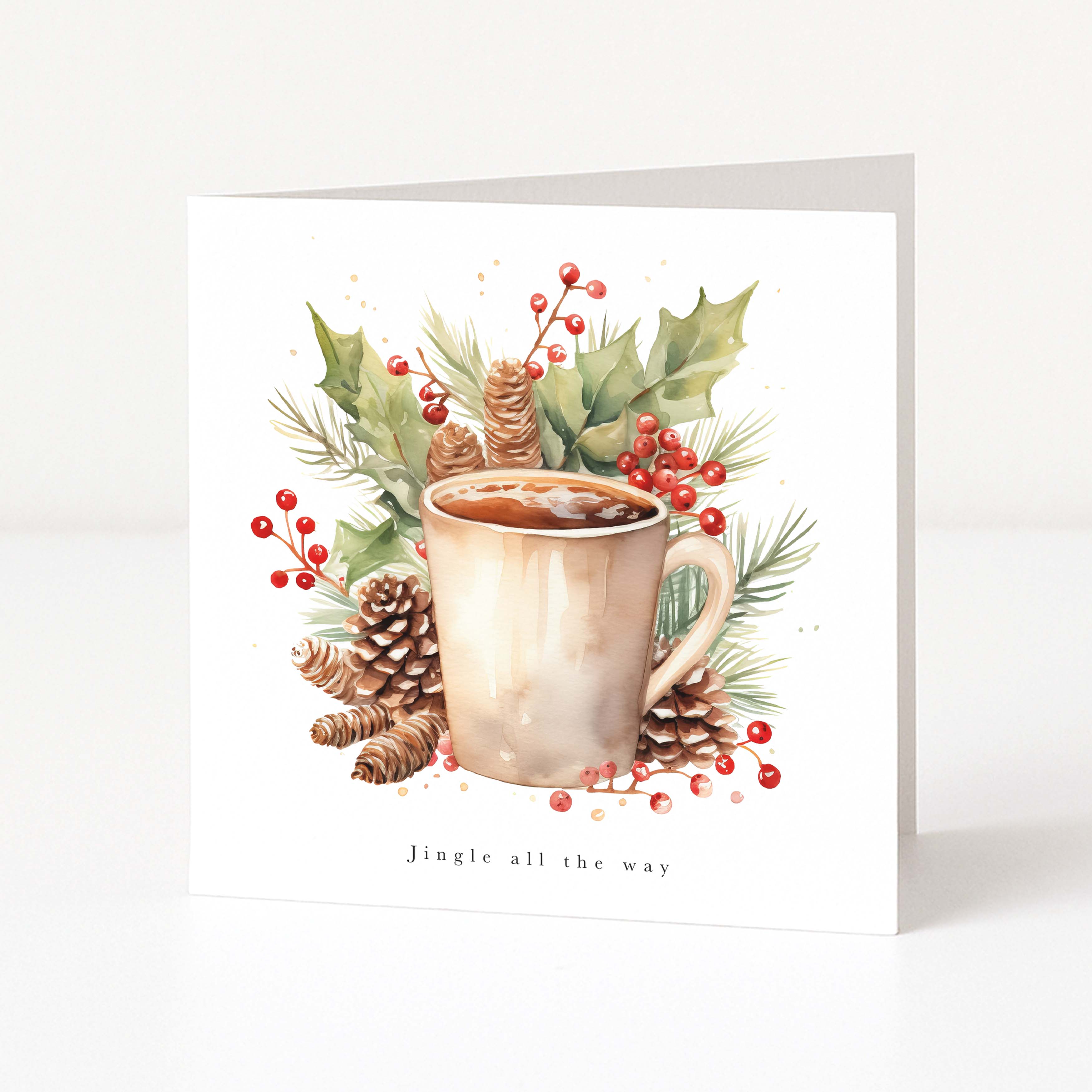 A square white Christmas greeting card standing upright on a light gray surface, featuring a watercolor-style illustration of a mug surrounded by pinecones, holly leaves, pine needles, and red berries, with the text 'Jingle all the way' at the bottom.
