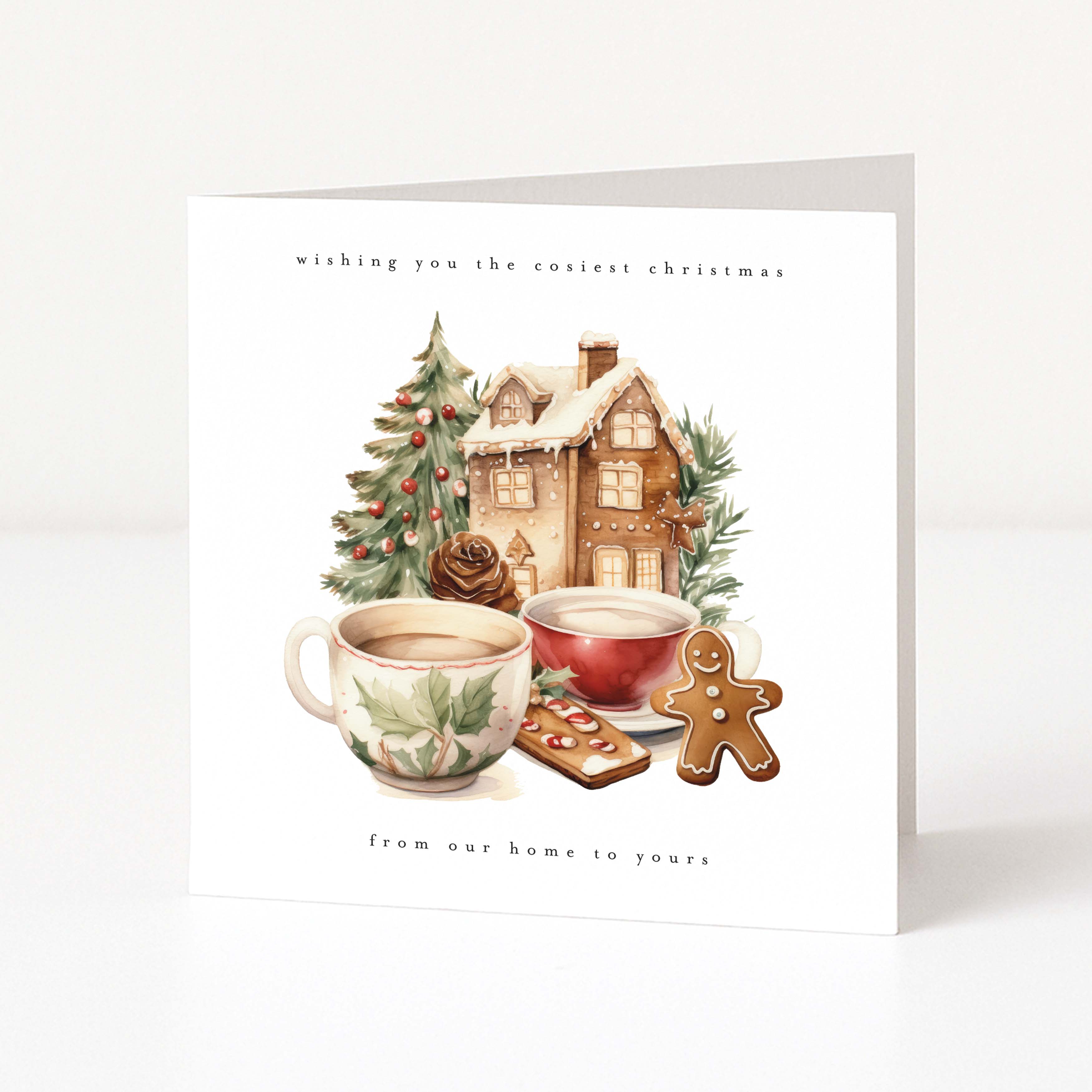 A square Christmas greeting card standing upright on a white surface against a light gray background. The card features a watercolor-style illustration of a cozy house, a decorated tree, mugs of tea, a gingerbread cookie, and the text 'wishing you the coldest Christmas' at the top and 'from our home to yours' at the bottom.