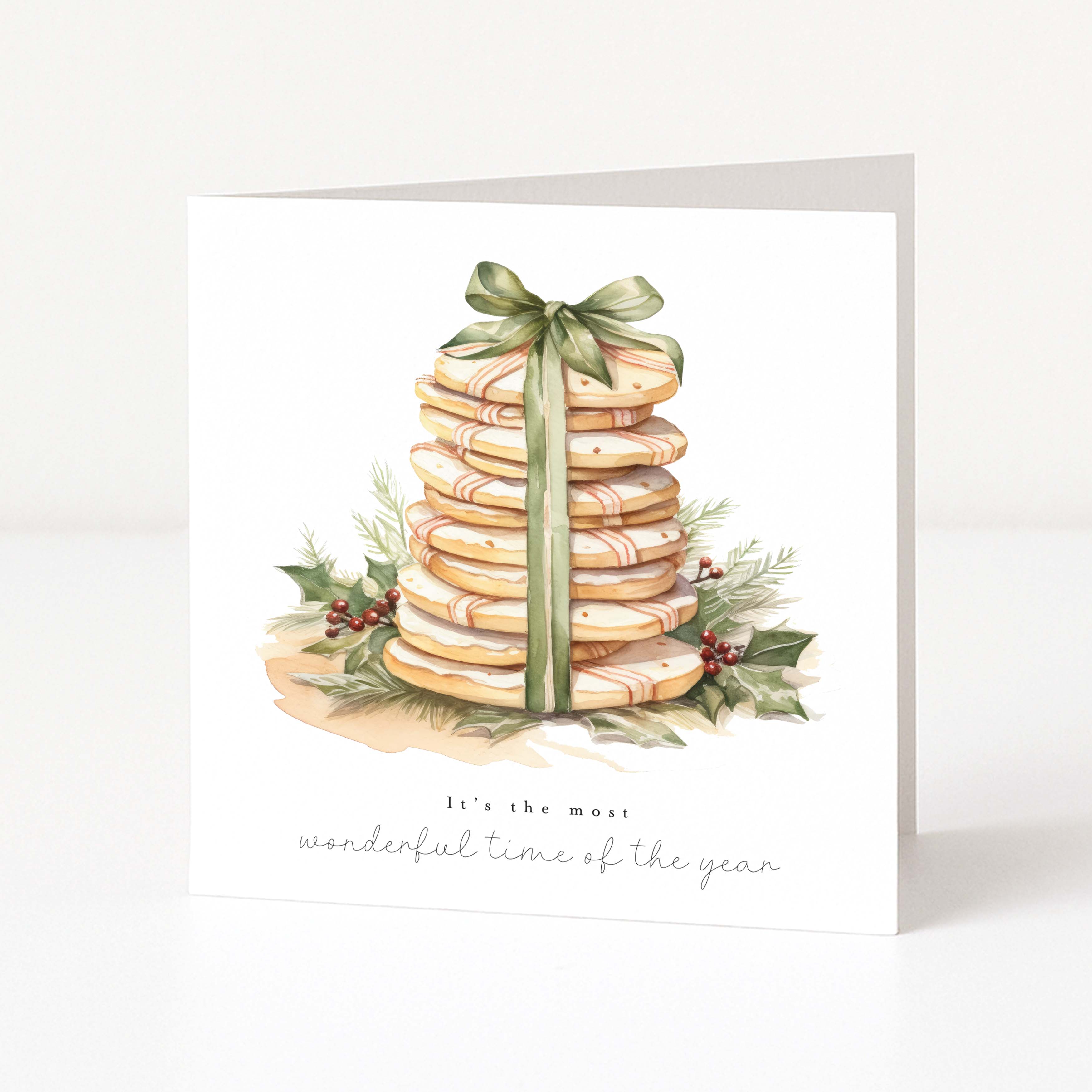A square white Christmas greeting card standing upright on a white surface, featuring a watercolour illustration of a stacked gift box with a green ribbon and holly, with the text 'It's the most wonderful time of the year' below.
