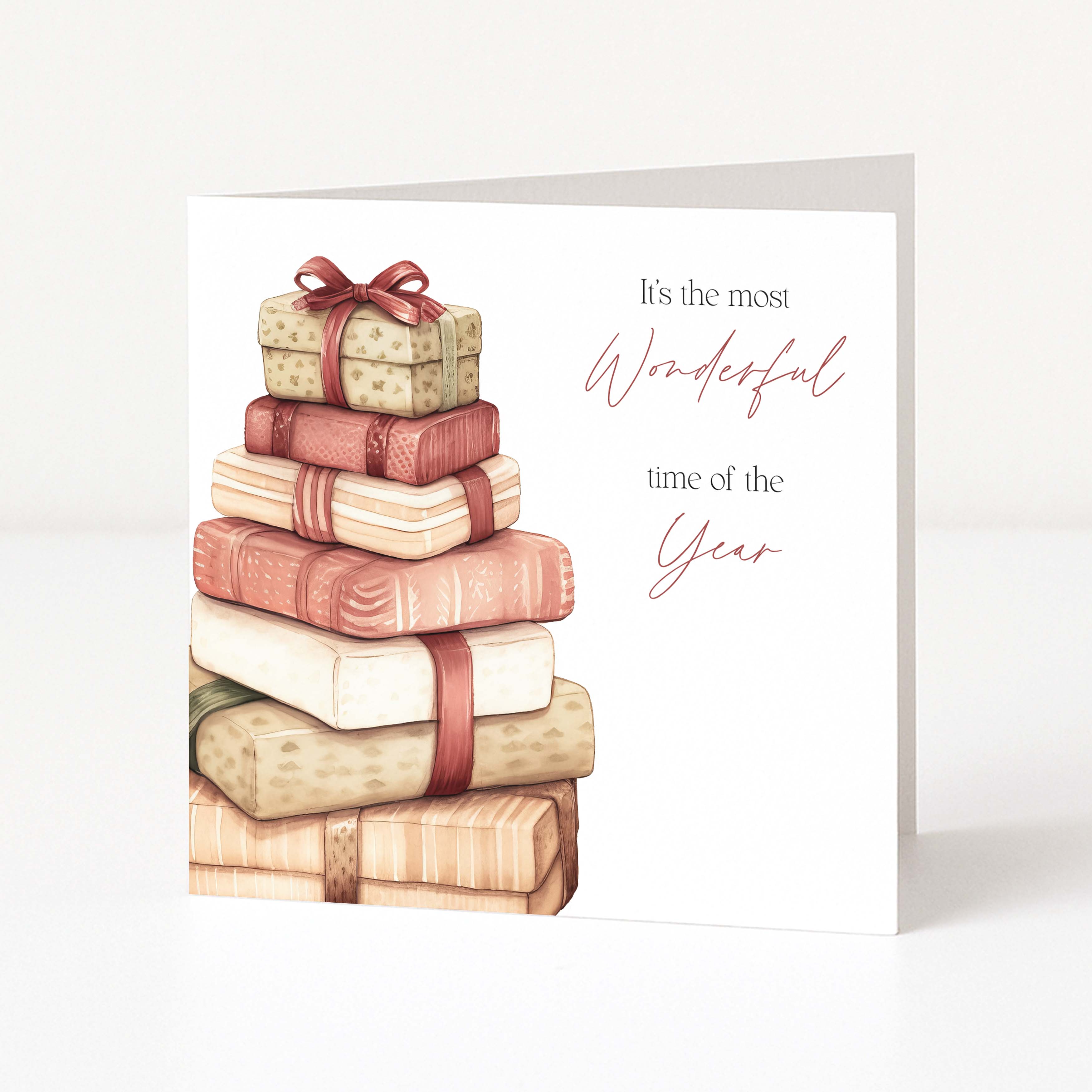 A white Christmas greeting card standing upright, featuring an illustration of stacked wrapped presents in soft beige, pink, and cream tones with a red ribbon. The right side of the card reads, 'It's the most Wonderful time of the Year' in black and dusty rose script.