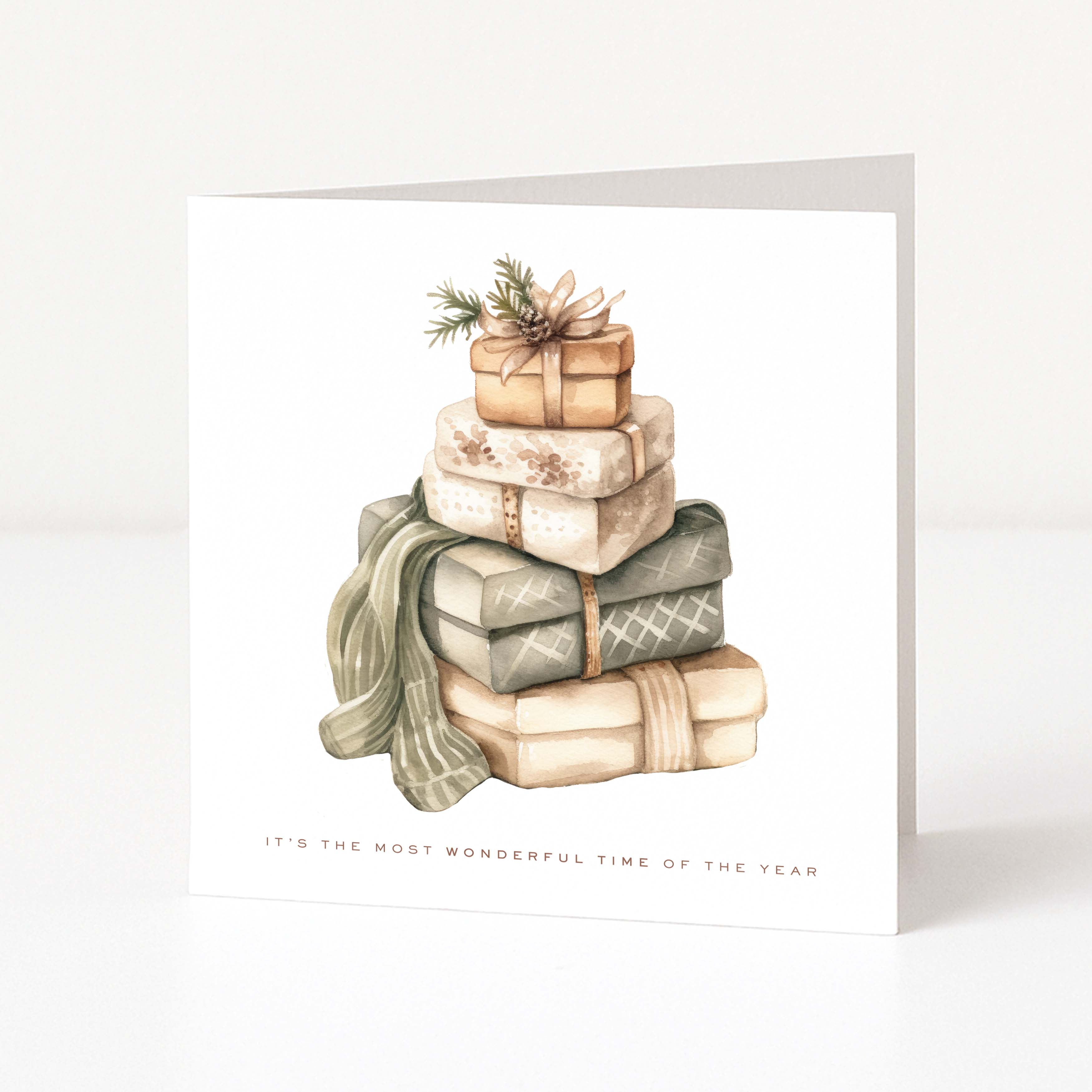 A square greeting card with a white background featuring a watercolour illustration of a stack of wrapped Christmas presents and a green scarf. The card reads 'IT'S THE MOST WONDERFUL TIME OF THE YEAR' at the bottom.