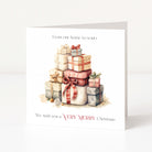 A square Christmas greeting card standing upright on a white surface against a light neutral background. The card features a watercolour illustration of stacked gift boxes with red, pink, beige, and gray wrapping, with the text 'From our home to yours' at the top and 'We wish you a VERY MERRY Christmas' at the bottom.