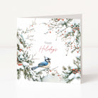 A square holiday greeting card standing upright on a white surface. The card features a winter illustration with snow-covered branches, red berries, and a blue jay perched near the lower centre, with the words 'Happy Holidays' printed in the middle.